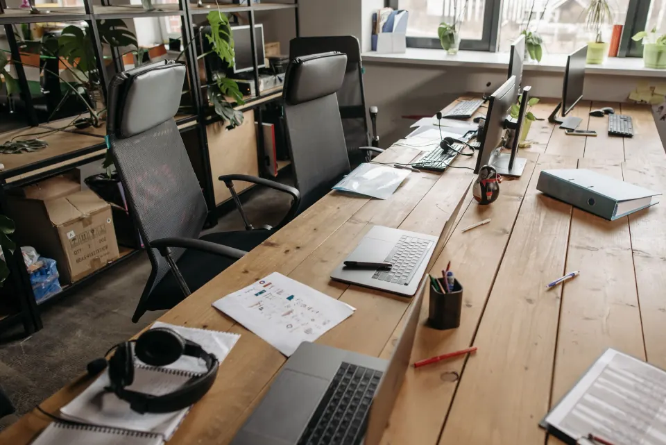 An open-plan office desk with laptops, monitors, headsets and printed charts