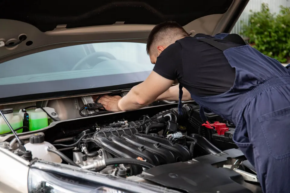 Mechanic in overalls reaching into a car's open engine bay to work on a wiring loom